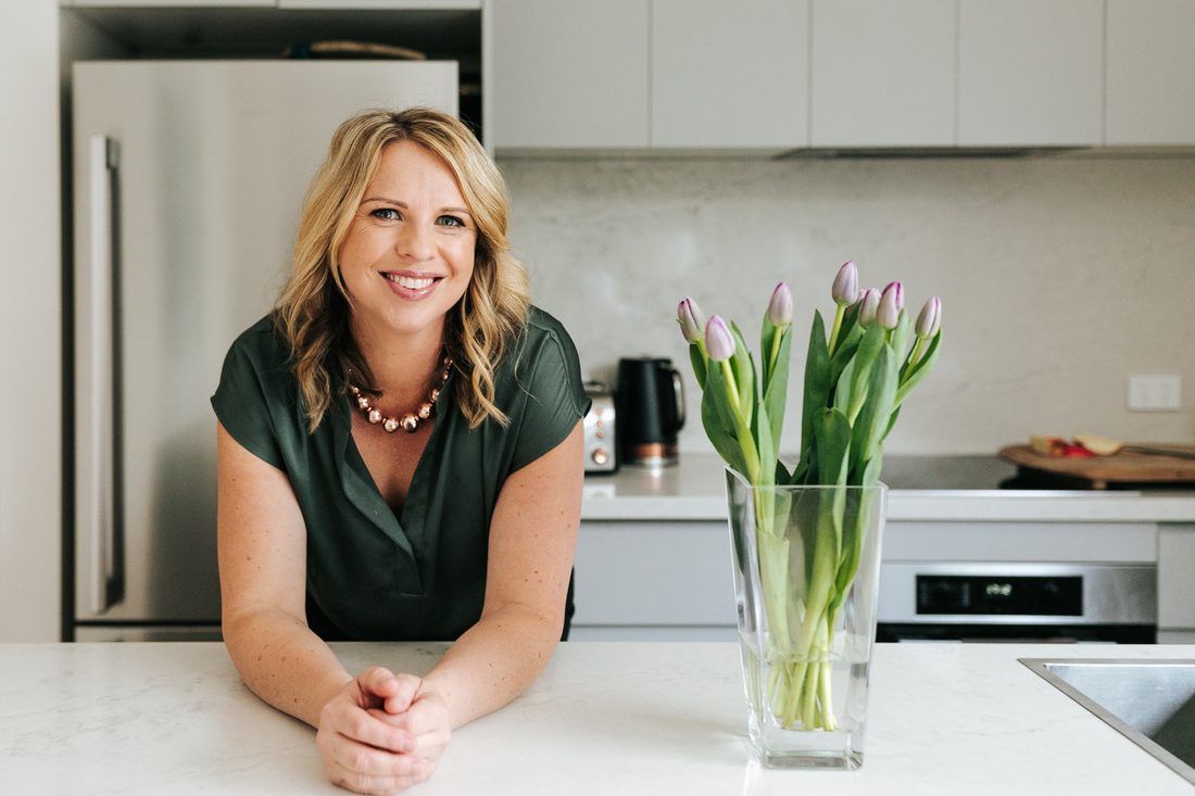 Georgie Beames leaning on her kitchen bench, smiling, with a vase of tulips beside her